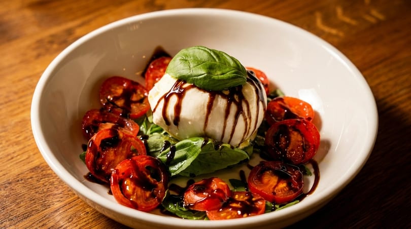 Burrata cheese with balsamic glaze, basil, and cherry tomatoes in a white bowl on a wooden table.